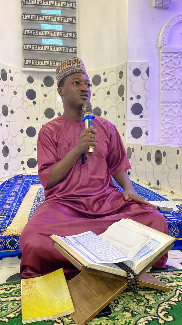 Alfacairo in maroon kaftan holding microphone with open Quran